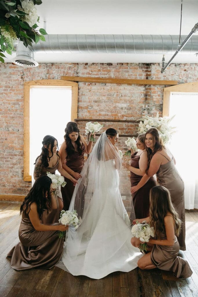 A bride in a fitted satin wedding gown with a long veil, surrounded by bridesmaids in champagne dresses